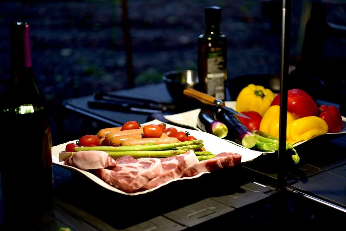 Assiette de charcuterie et légumes frais sur table de camping en soirée, avec bouteilles de vin et fruits à proximité